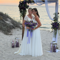 Chantal en Noa in trouwjurken onder een boog met paarse bloemen op het strand van Kos bij zonsondergang.