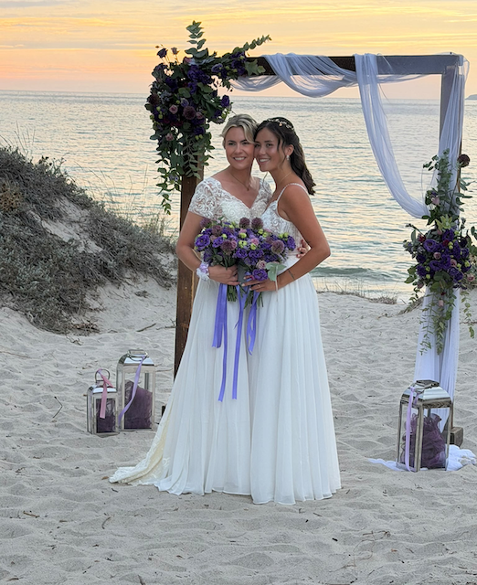 Chantal en Noa in trouwjurken onder een boog met paarse bloemen op het strand van Kos bij zonsondergang.