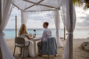 Bruidspaar geniet van een romantisch privédiner onder een witte cabana op het strand bij Karakter op Curaçao, met uitzicht op zee.